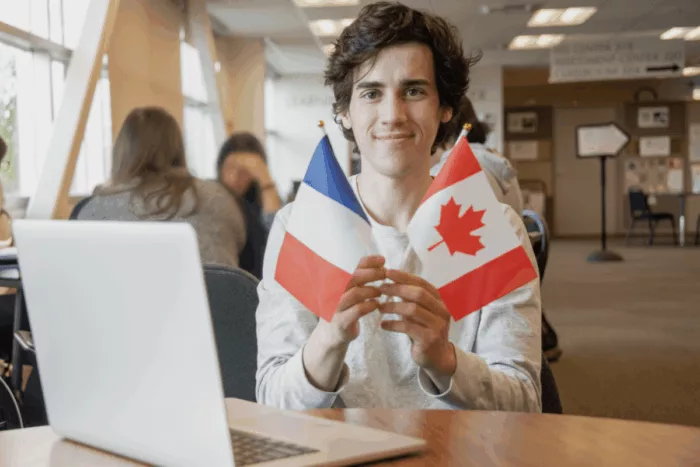 Étudiant en management international, souriant dans une bibliothèque et tenant à la main un drapeau français et un drapeau canadien. Un ordinateur portable est ouvert devant lui.